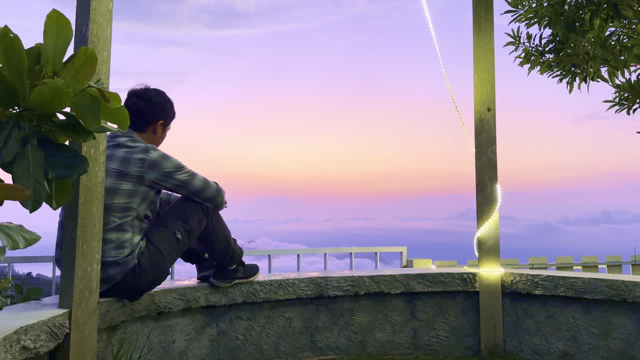 An adult man sits alone and lowers his head and rests on his knees. View of the morning sunrise sky.
