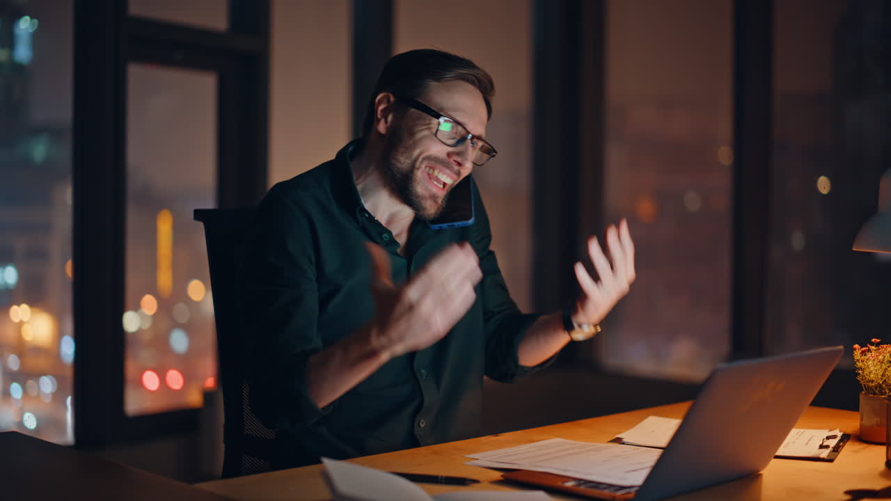 Investitore impegnato che parla al cellulare e tiene in mano dei documenti in un ufficio di notte, primo piano
