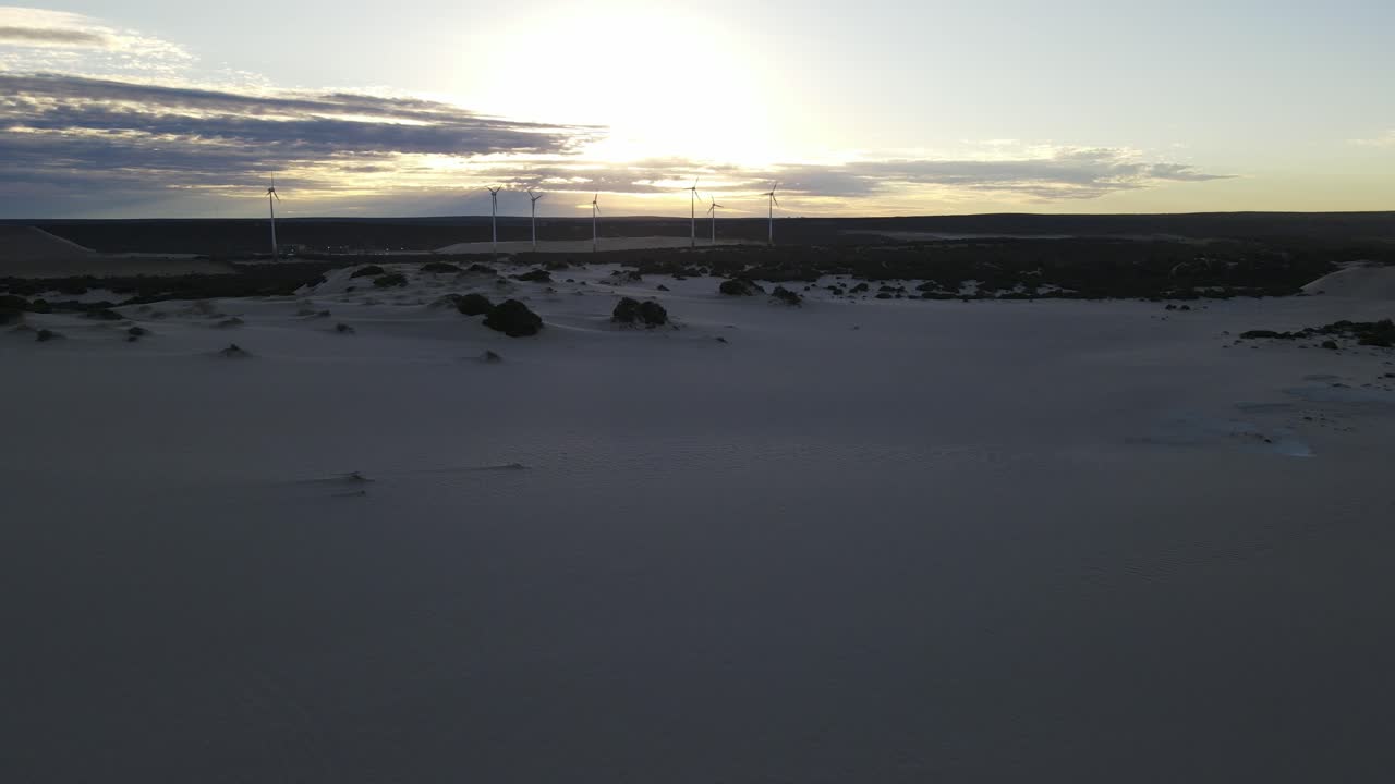 Drone aerial moving left over sand dunes showing a sunrise by some wind mills