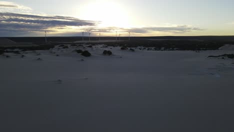 Drone-aerial-moving-left-over-sand-dunes-showing-a-sunrise-by-some-wind-mills