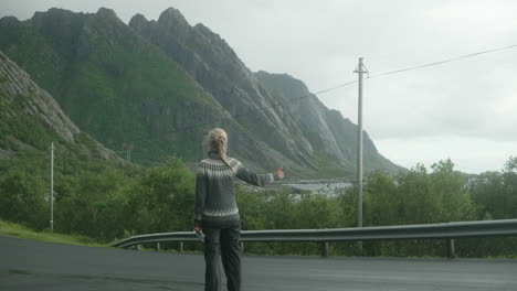 Pretty-Blonde-Girl-Hitchhiking-at-the-Side-of-the-Road-in-the-Lofoten-Islands-on-a-Cold-Cloudy-Rainy-Day,-Beautiful-Rocky-Mountains-in-the-Background,-Cars-Are-Passing,-Norway