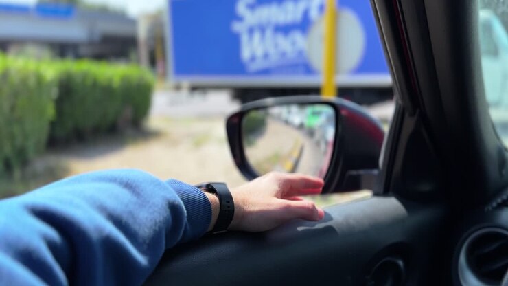 Inquadratura soggettiva dall'interno di un'auto parcheggiata che mostra la mano di un conducente appoggiata sulla portiera vicino allo specchietto laterale, con strada urbana, traffico e vegetazione visibili alla luce del giorno.