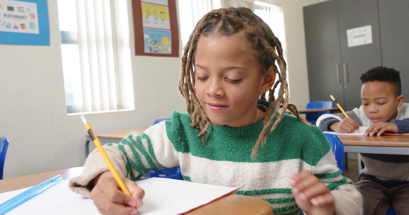 Bambini afroamericani, un bambino con le trecce che prende una matita gialla e scrive per finire il lavoro alla scrivania.