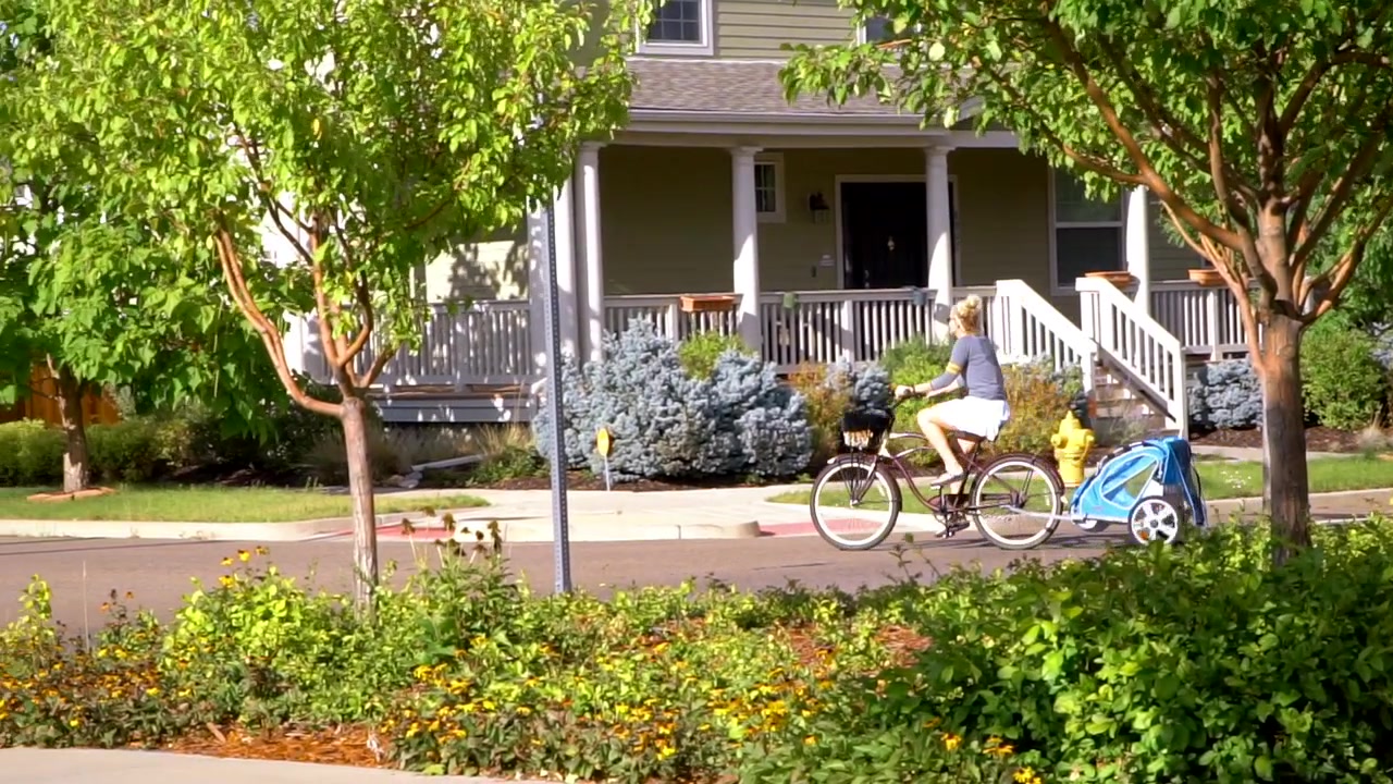 スローモーション。コロラド州ステープルトンにて、自転車用トレーラーで子供を牽引しながら近所を走る母親