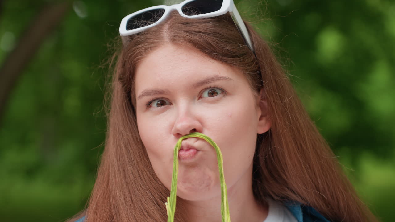 Close up of funny student holding green thread under nose pretending mustache while turning eyes sideways and reacting playfully as thread slips off, sitting on park bench