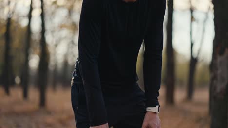 Close-up-footage-of-a-brunette-man-in-a-black-sports-uniform-running-up-to-his-destination-in-the-autumn-forest-and-taking-a-short-break-wiping-away-sweat-during-his-morning-jog-in-a-sunny-autumn-forest