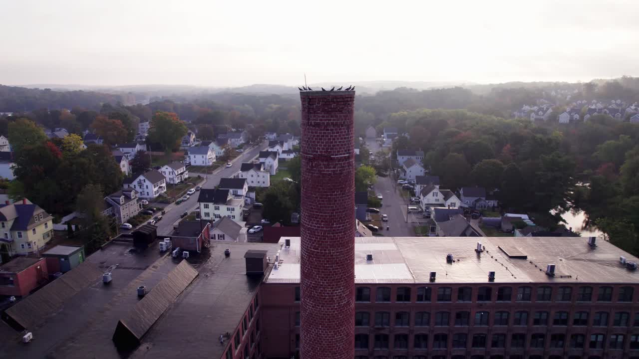 Drone approaching a brick mill smokestack with a group of birds perched at the top. Overlooking residential areas