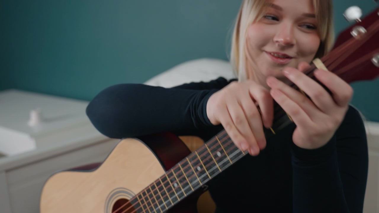 Donna bionda in un nuovo appartamento accorda la chitarra, con scatoloni da disfare e un letto vicino a un comodino bianco. Prova concentrata di fingerstyle e delicato strimpellio, mentre si ambienta nella sua nuova casa con una calma luce diurna e un'atmosfera di speranza.