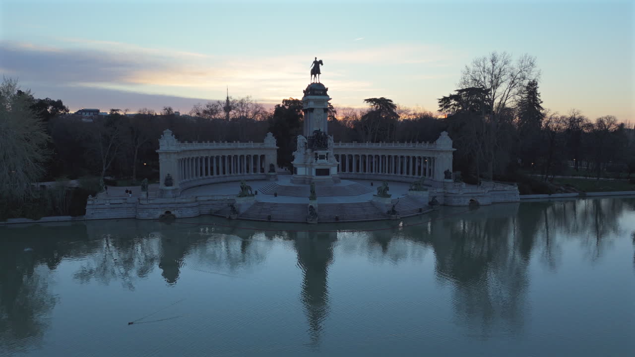 Drone flying low over Retiro pond reflecting Alfonso XII monument at sunrise, showcasing statues, lake, stunning gardens, historical architecture, trees, and tranquil morning in iconic city park