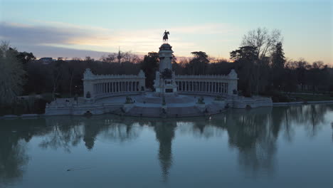 drone flying low over retiro pond reflecting alfonso xii monument at sunrise, showcasing statues, lake, stunning gardens, historical architecture, trees, and tranquil morning in iconic city park