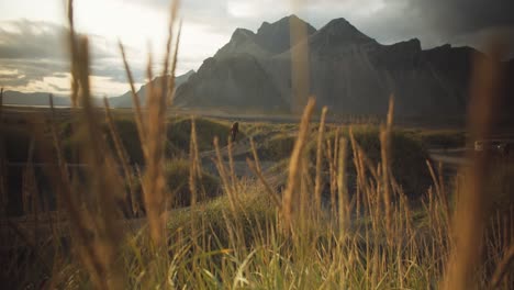 Wanderer-Geht-Auf-Einer-Kleinen-Düne-Mit-Schwarzem-Sand-In-Der-Ferne-Einer-Grasbewachsenen-Berglandschaft-Spazieren