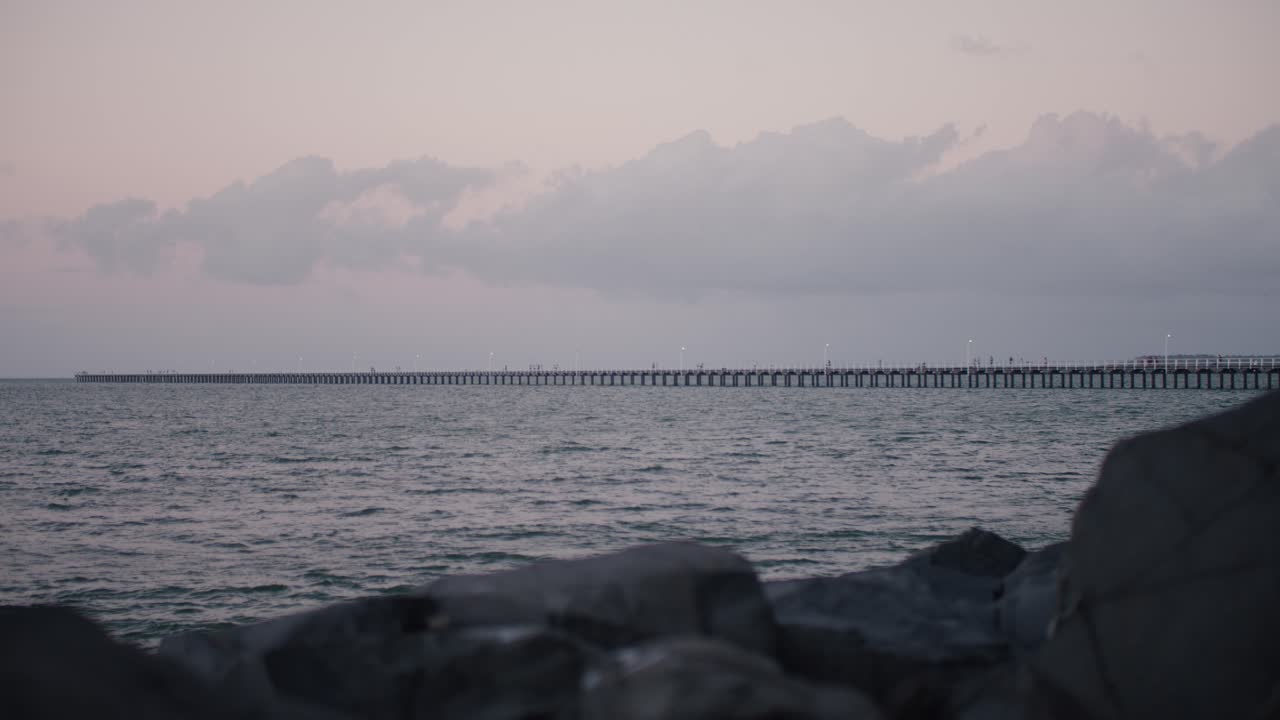 매우 긴 부두는 해가 질 때 해변의 제방에서 광활한 바다를 향해 뻗어 있으며 오후 하늘에 분홍색과 파란색 파스텔 색상을 남깁니다.
