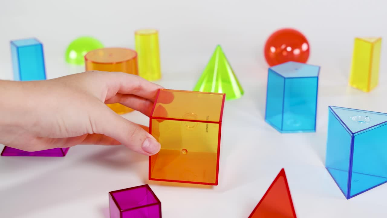 Hands interact with colorful 3D shapes on a white surface, demonstrating basic geometry concepts in a playful setting