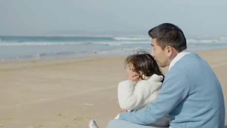 Japanischer-Vater-Und-Süße-Kleine-Tochter-Sitzen-Am-Meer,-Reden-Und-Genießen-Die-Aussicht