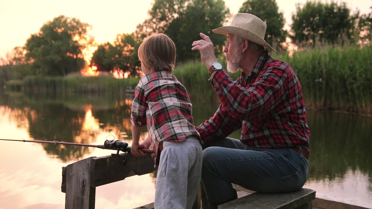 Side view of a grandfather and his grandson fishing sitting on the lake pier on a summer day at sunset