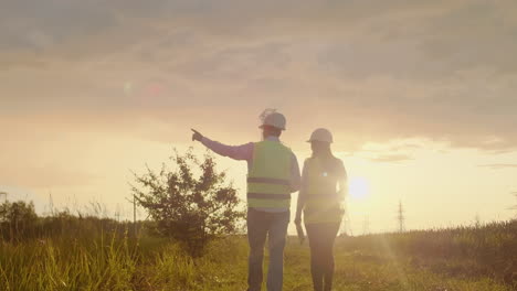 la vista desde la parte trasera: dos ingenieros un hombre y una mujer con cascos con una tableta de ingeniero caminan en el campo con torres de electricidad y discuten la construcción adicional de torres