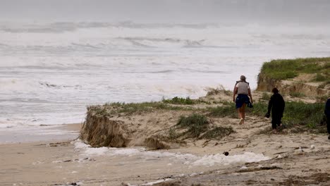 two individuals walk along an eroded coastline, observing the effects of seafoam and erosion on the pacific ocean shore cyclone alfred