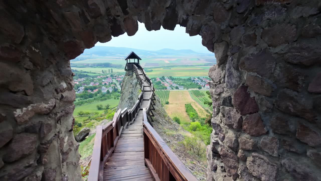 Dolly-out view through a stone archway at Boldogkőváralja Castle, Hungary, revealing a wooden walkway, medieval architecture, and a panoramic countryside of fields, hills, and greenery in daylight
