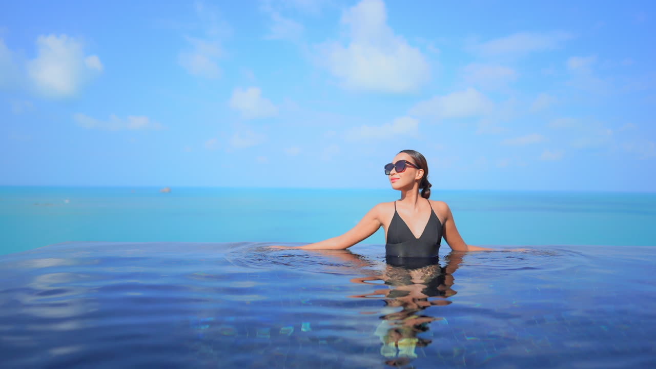 Confident exotic female in a black swimsuit in infinity pool with a tropical sea horizon in background, luxury vacation concept, full frame