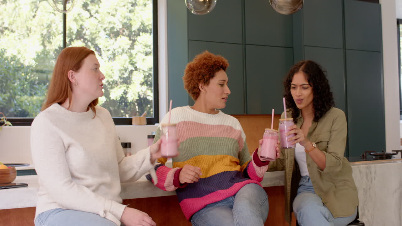 Drinking smoothies, diverse female friends hanging out and chatting in kitchen