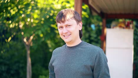 Close-up-Portrait-Young-man-stands-against-backdrop-of-summer-house-and-green-trees-and-smiles