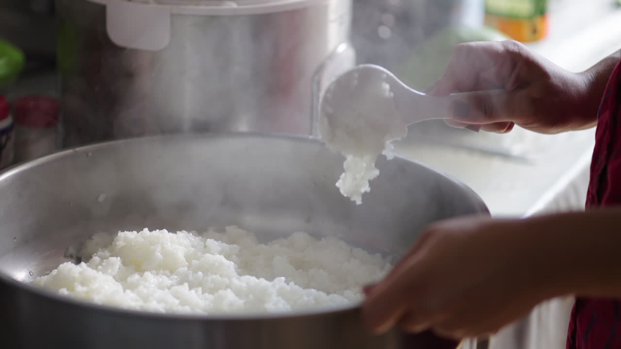Cook uses paddle to fold and aerate steaming short‑grain rice in wide metal pot, plump translucent grains releasing wisps of steam as gently fluffed for sushi or rice bowl in professional kitchen