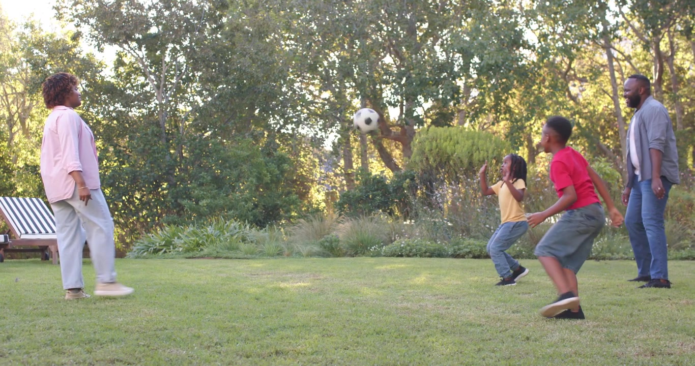African American family playing soccer on yard, man moving ball, kids chasing, senior woman kicking