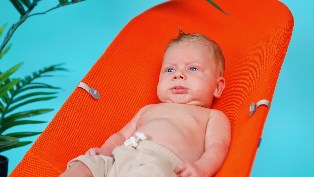 Blue-eyed blond Caucasian infant lies in the orange chair. Portrait of a baby resting. Blue backdrop.