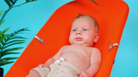 blue-eyed blond caucasian infant lies in the orange chair. portrait of a baby resting. blue backdrop.