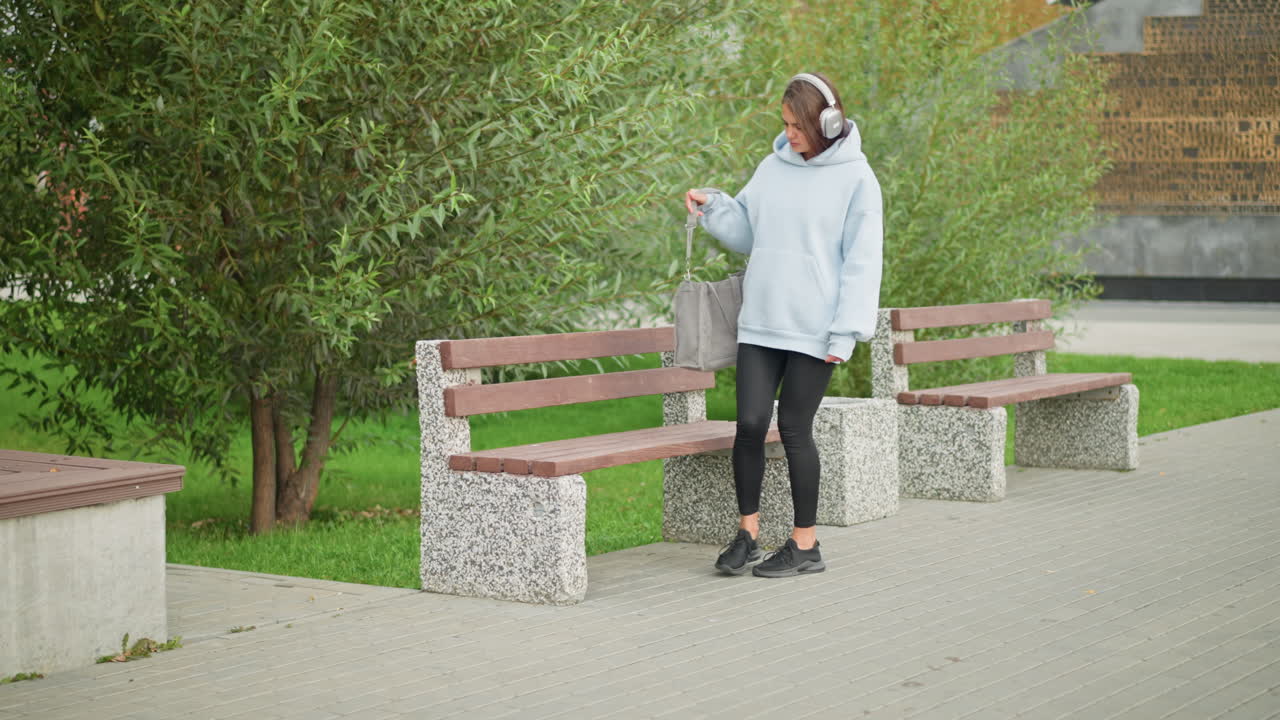 Pretty girl listening to music with headphones, walking slowly to concrete bench, sitting, and trying to open handbag, background includes lush green trees and a peaceful park atmosphere