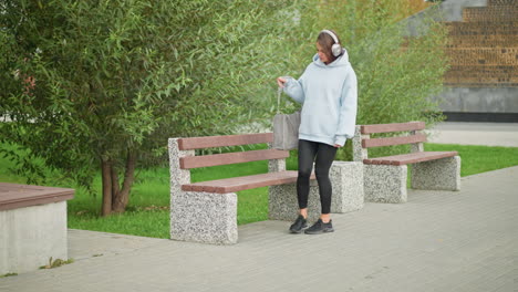 pretty girl listening to music with headphones, walking slowly to concrete bench, sitting, and trying to open handbag, background includes lush green trees and a peaceful park atmosphere