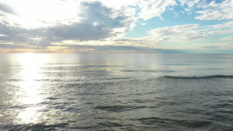 AERIAL:-Flying-Backwards-with-Calm-and-Peaceful-Sea-with-Reflection-of-Sky-and-Cloudy-Sky-in-the-Background-with-Sandy-Beach