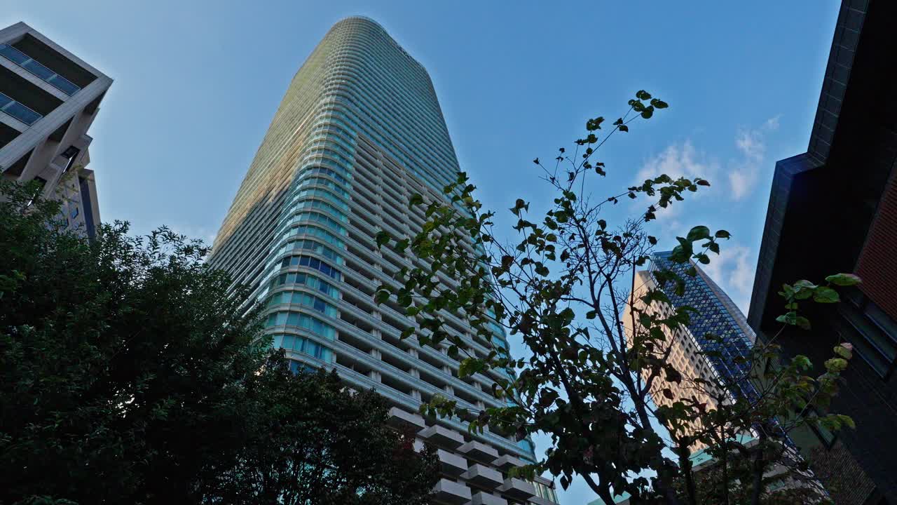 An upward view of a sleek, modern Tokyo skyscraper, its curved lines and glass rising impressively against a clear blue sky, framed by urban trees.