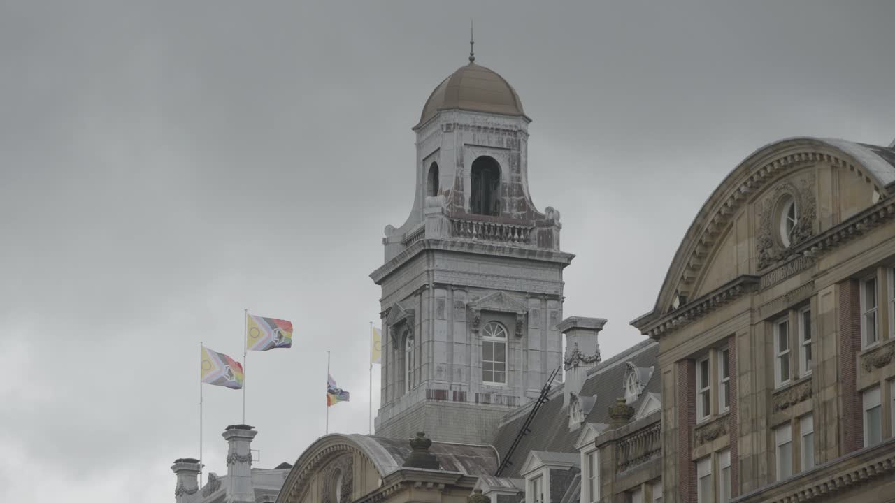 Pride and LGBTQ+ inclusive flags waving in the wind on top of a classical building, shot on a cloudy day.