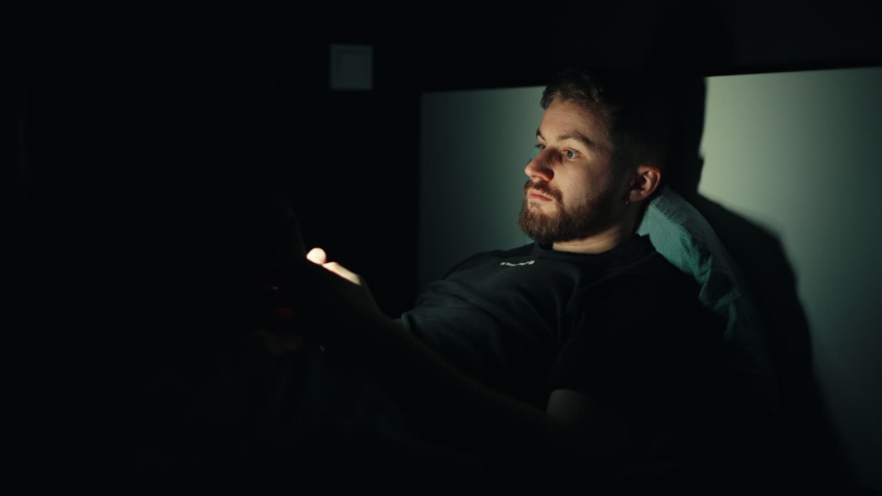 Un giovane uomo sdraiato al buio, con gli occhi illuminati dallo schermo del telefono mentre scorre all'infinito. Uno sguardo cinematografico alle abitudini notturne davanti allo schermo e alla dipendenza digitale prima di dormire.