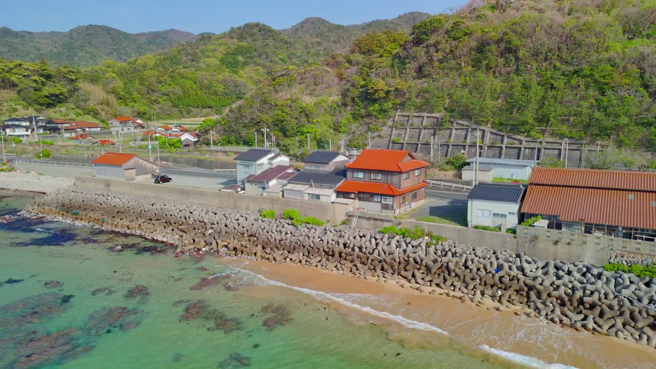 An aerial shot pans right along a scenic Japanese coastline, passing a village with a sea wall and revealing a road tunnel nestled in the lush mountains