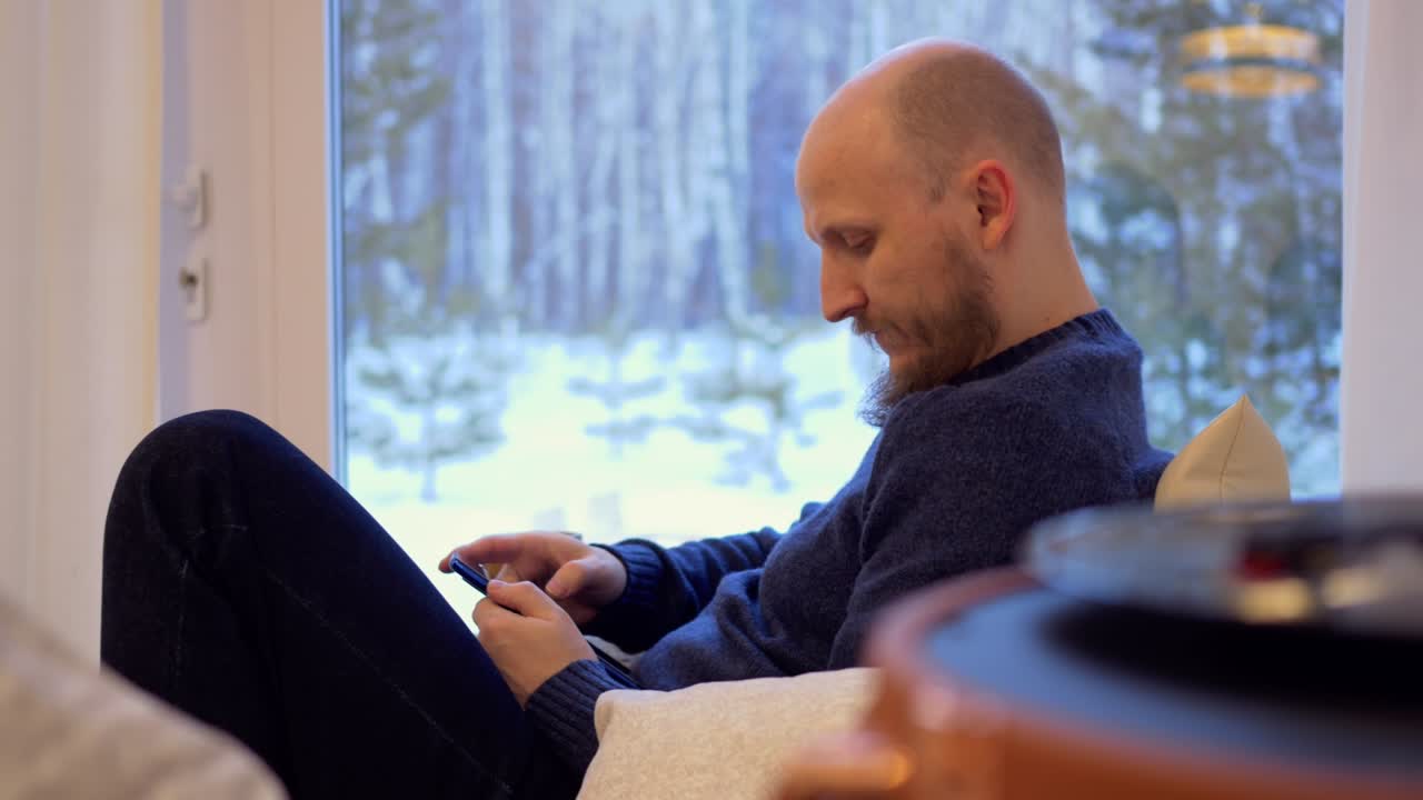 수염이 있는 남자가 소파에 앉아서 전화로 문자를 확인하고, 재미를 위해 친구들과, 일하기 위해 동료들과 소통하기 위해 스마트폰을 사용한다.