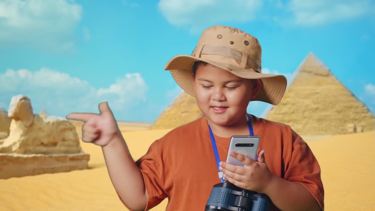 Asian Boy With A Hat And Binoculars Looking At Smartphone Then Smiling And Pointing To Side While Traveling In Giza Pyramid. Boy Researcher Examines Something, Travel Adventure, Close Up