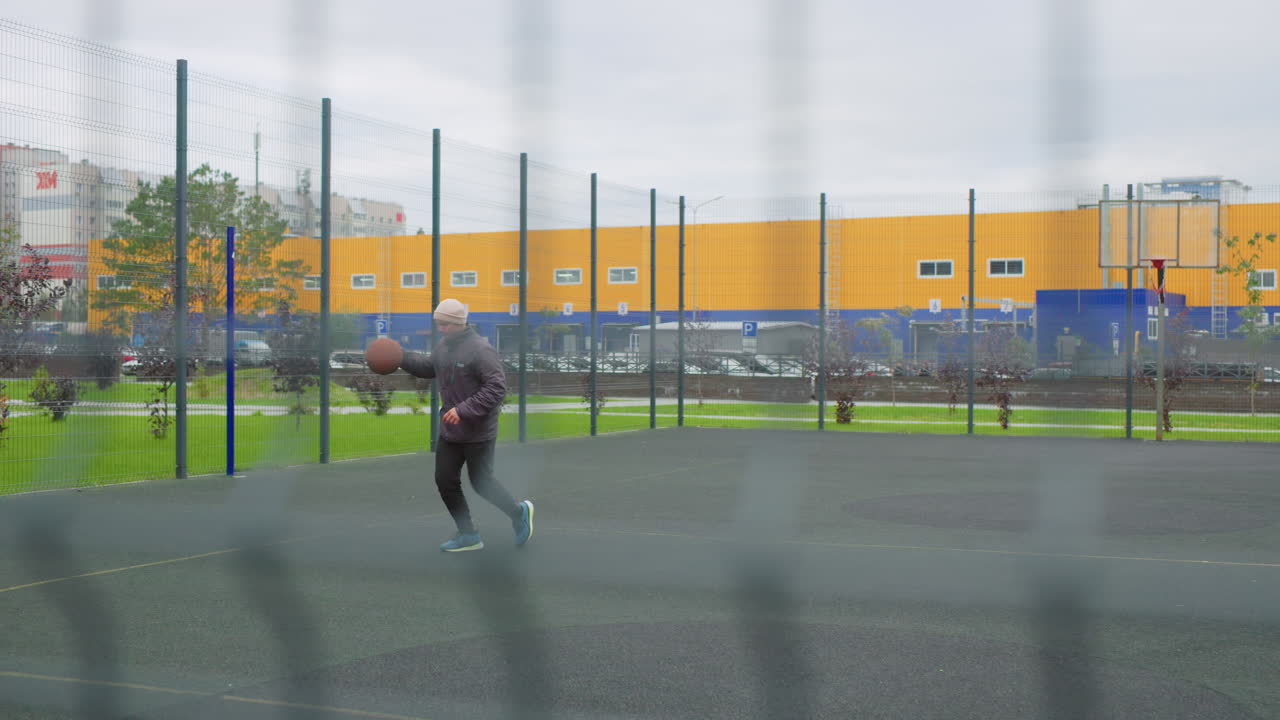 outdoor urban basketball court, player shoots into rim, fenced court with yellow warehouse backdrop, cloudy sky and wet pavement, hoodie and sneakers, focused warmup dribble and jump, closeup