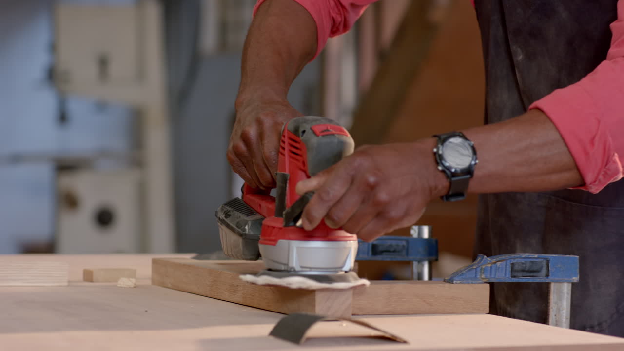 Senior African American man operating power tool on wood board in woodshop, with apron, copy space