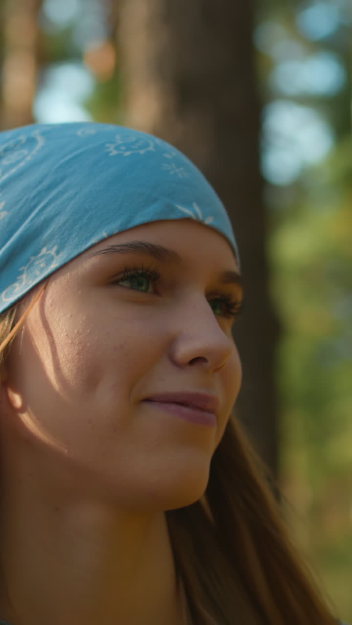 una mujer con un pañuelo azul y una sonrisa cálida camina a través de un bosque iluminado por el sol, sus hoyuelos visibles mientras mira satisfecha a su alrededor, la luz del sol se filtra suavemente a través de los árboles altos