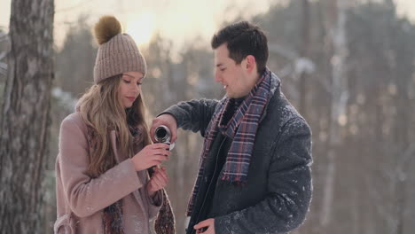 a loving couple man and woman in the winter forest drinking tea from a thermos. stylish man and woman in a coat in the park in winter for a walk. slow motion