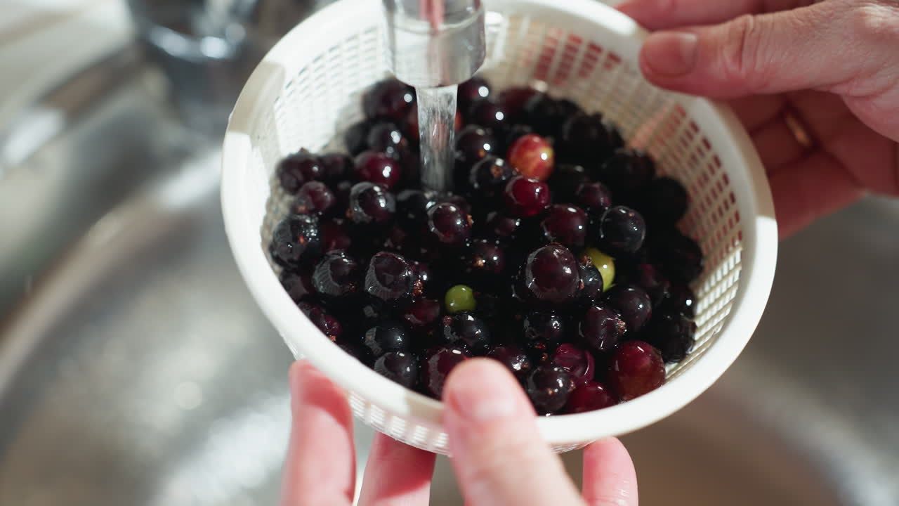 Close up of hand holding basket of dark ripe berries under running tap water in kitchen sink, rinsing fruit thoroughly, person wears simple gold ring, light reflects on wet surface of berries