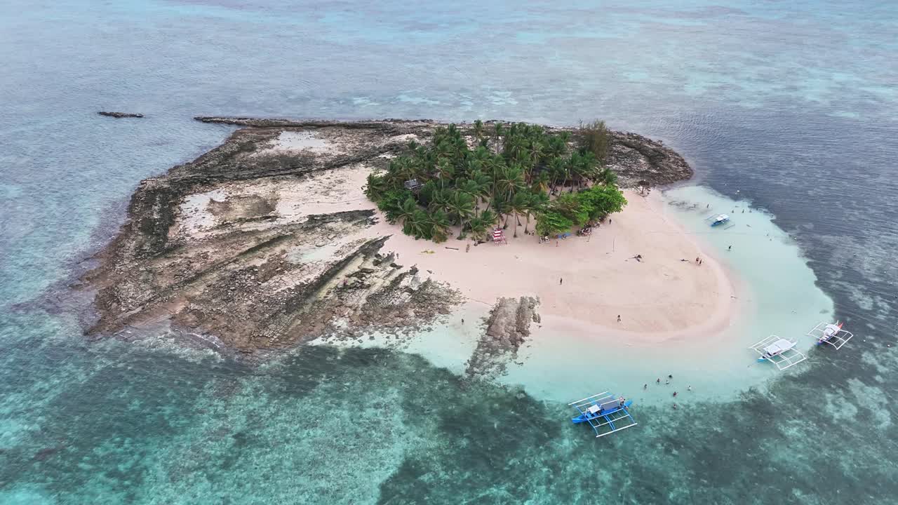 Aerial perspective of Guyam Island in Siargao, Philippines, revealing its palm-filled core, sand shores, coral formations, and turquoise waters with traditional boats and swimmers surrounding island