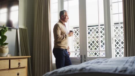 Happy-caucasian-man-looking-through-window-and-drinking-coffee-in-bedroom