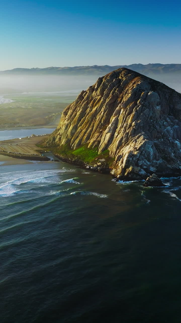 Flying closer to a pyramid-shaped mountain at the coast of Pacific. Little white waves coming slowly to the shore. White fog covering the horizon. Vertical video