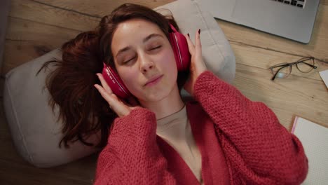 Top-view-of-a-confident-brunette-girl-in-wireless-red-headphones-and-a-sweater-listening-to-music-during-her-break-between-studying-and-self-development-in-a-modern-apartment-on-the-floor