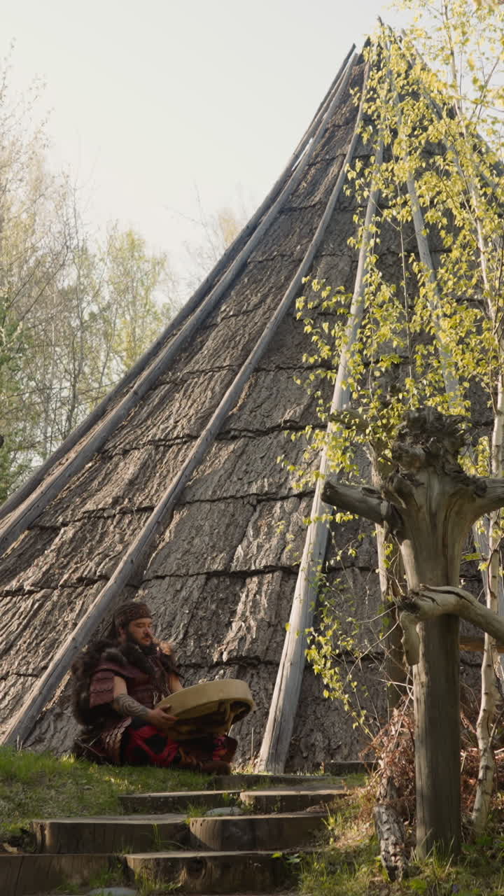 Musicain in Siberian shaman costume plays ritual drum sitting near ail building. Reconstruction of ancient Altai village on spring day