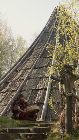 musicain in siberian shaman costume plays ritual drum sitting near ail building. reconstruction of ancient altai village on spring day