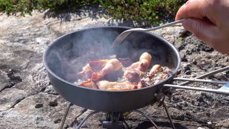 Person-cooking-bacon-in-camping-skillet-over-small-white-gas-burner